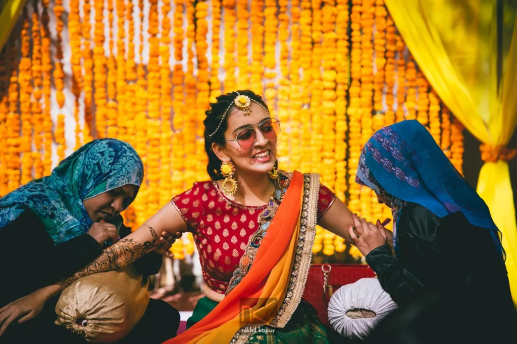 A bride laughing as her mehendi is applied, in front of a wall of marigolds